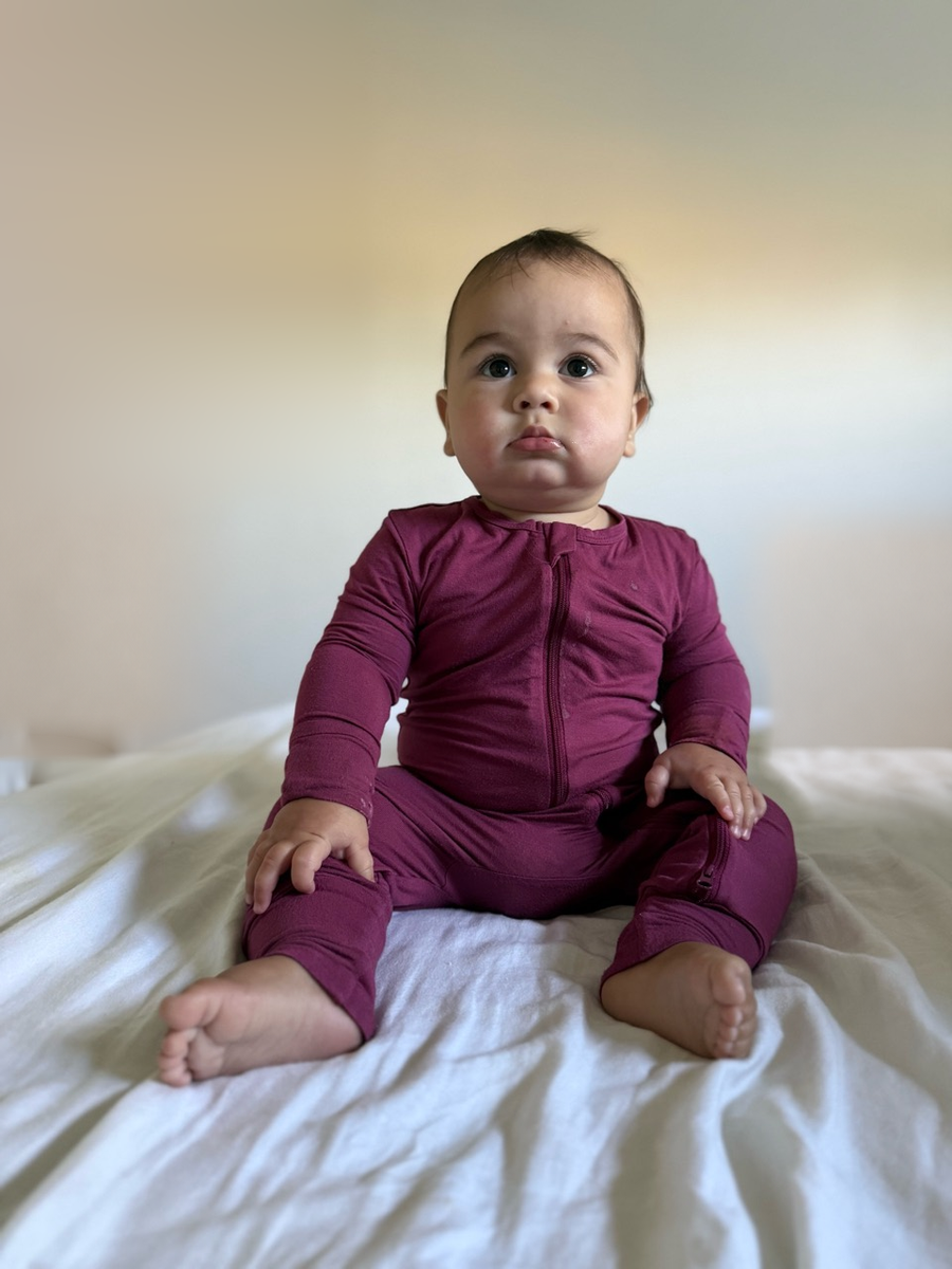 Jordan wearing the Berry Rose Garden long-sleeve two-way zip romper, seated on light bedding.