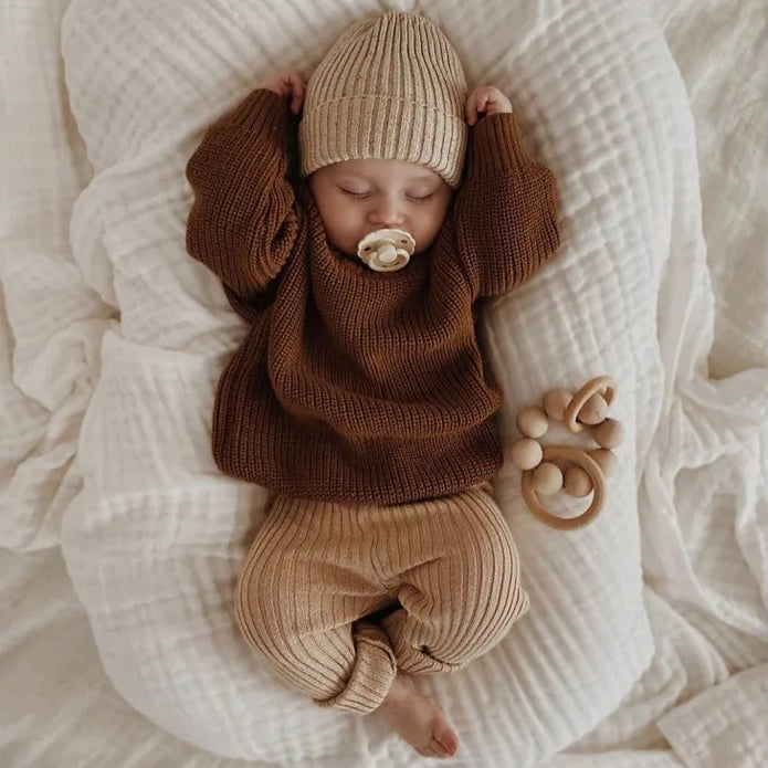Baby in brown outfit and hat lying on a white blanket with a wooden toy.