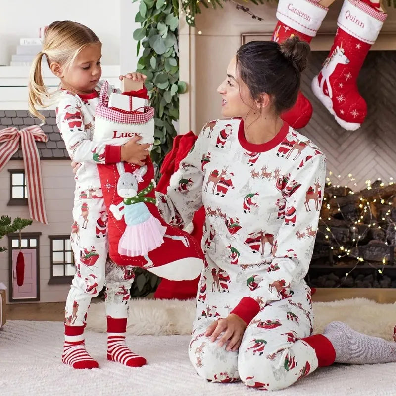 An adult and child wearing cream long-sleeve pajamas printed with Santa and reindeer, with red necklines and cuffs.