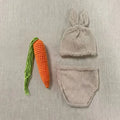 Beige knit baby rabbit hat with ears, knit diaper cover, and orange knit carrot prop laid flat on beige background