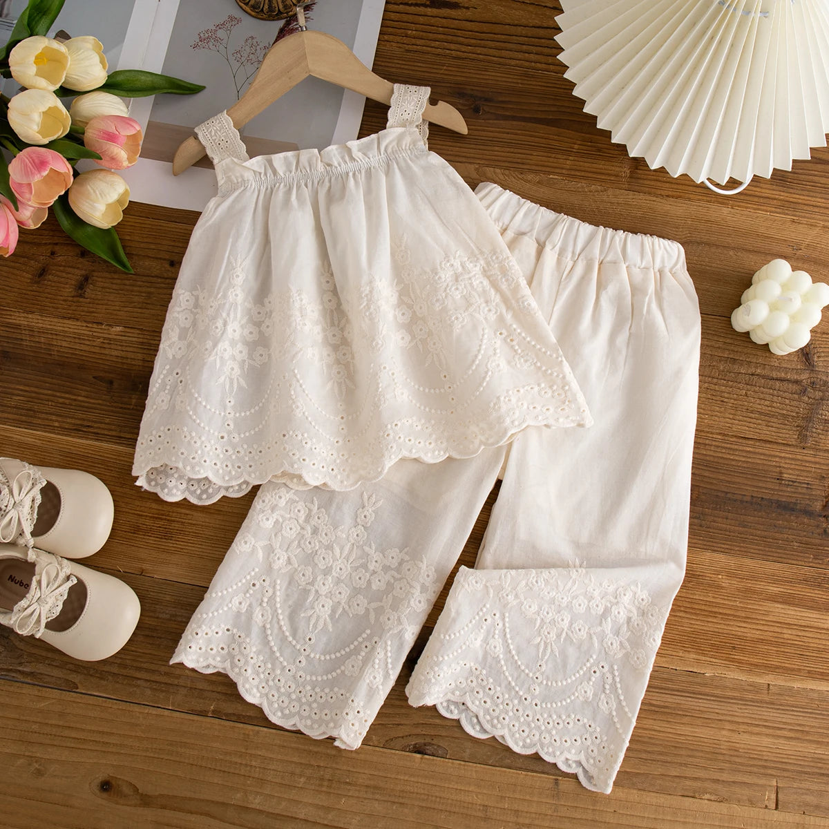 White lace baby outfit on a wooden surface with flowers and shoes.