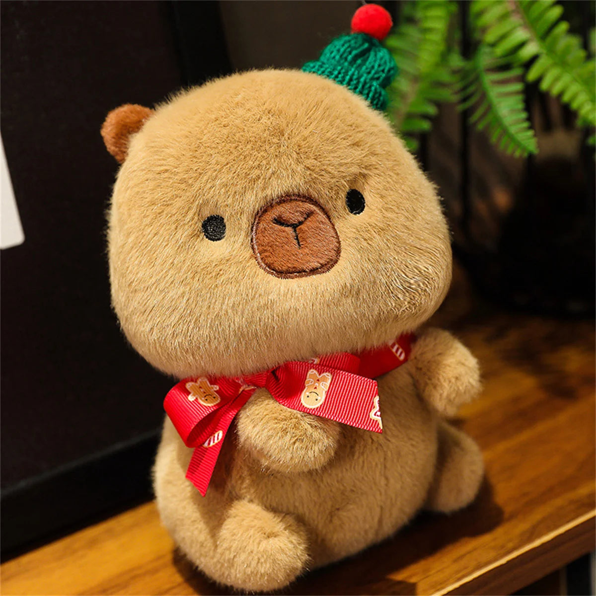 Brown capybara plush figure wearing a green pom-pom hat and red printed ribbon bow.
