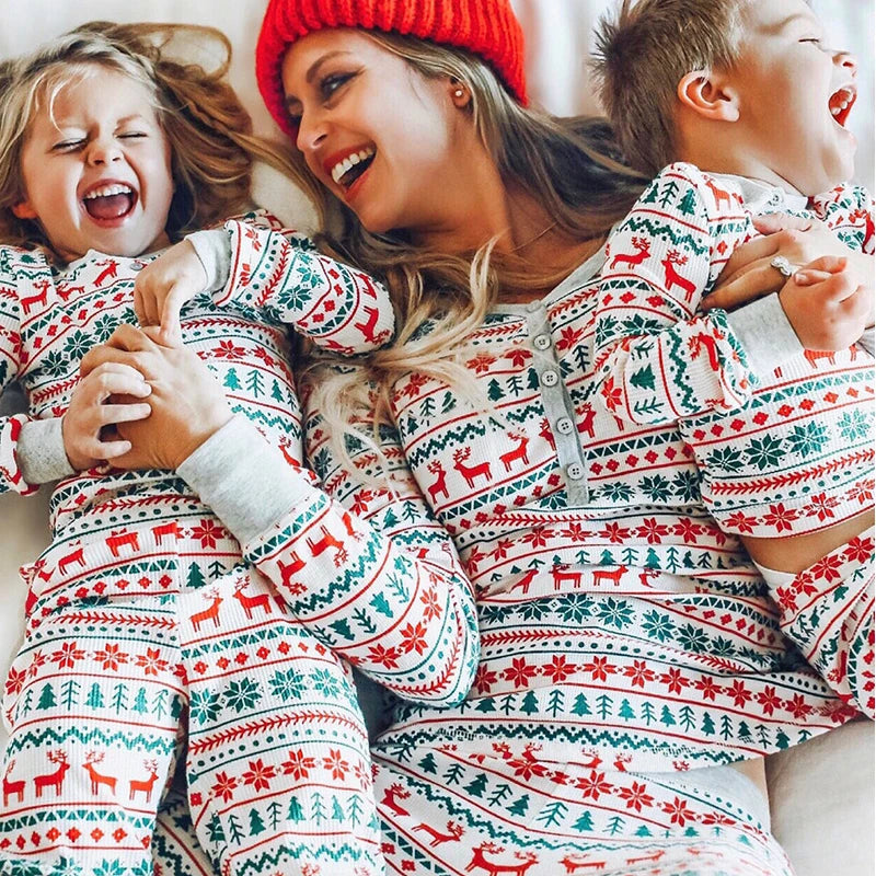 An adult and two children wearing white pajamas patterned with red reindeer, green trees, snowflakes, and geometric bands.