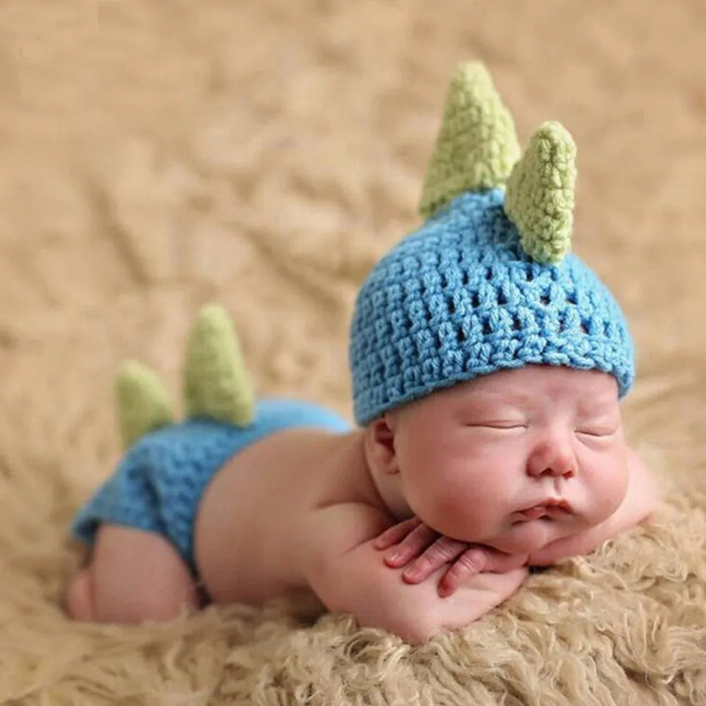 Baby wearing blue knitted dinosaur hat and diaper cover with green spikes