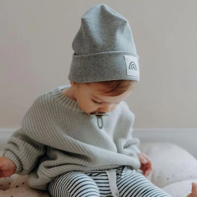 Child wearing a gray knit poncho and striped pants with a white hat featuring a rainbow emblem.