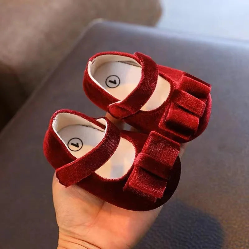Red Mary Jane baby shoes with instep straps and large folded bows across the toes, held in one hand.