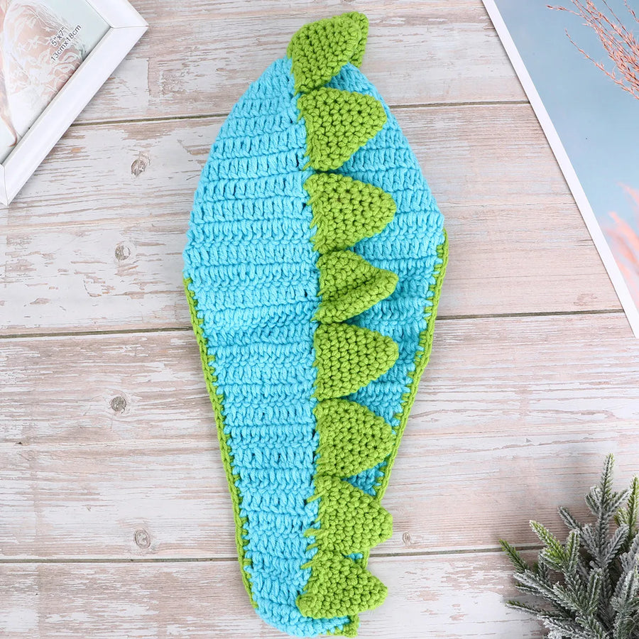 Blue crochet dinosaur hat with green spikes on wooden surface