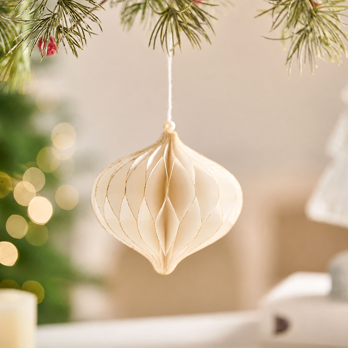 Cream honeycomb paper pointed bauble ornament with gold-colored edges, hanging from a cord on a pine branch.